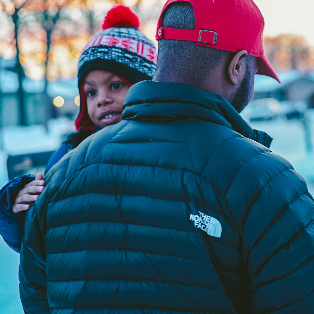 A child wearing a knit hat is being carried by an adult in a red cap and dark jacket, outdoors in a snowy setting.